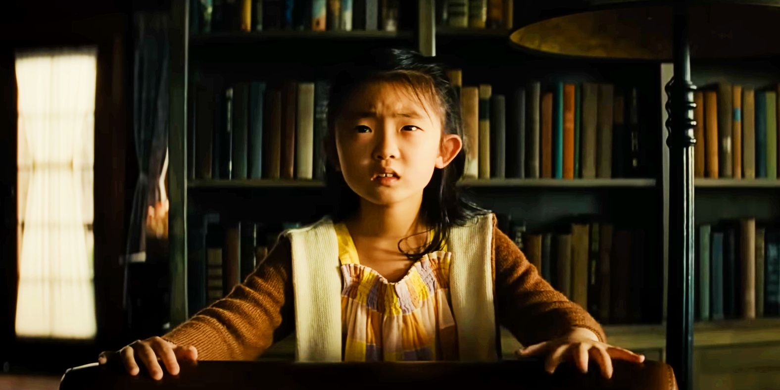 Knock at the Cabin'de kitap rafının önünde korkmuş görünen Wen rolünde Kristen Cui