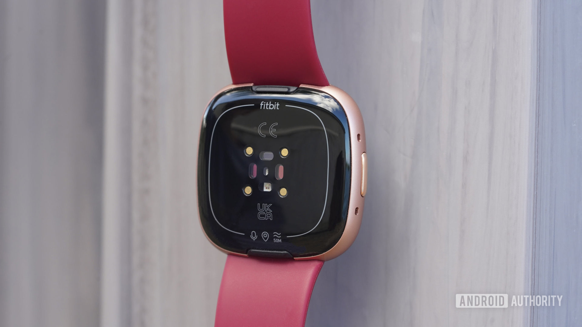 Gri bir yüzeyin önünde asılı duran bir Fitbit Versa 4, cihazın sensörlerini gösteriyor.