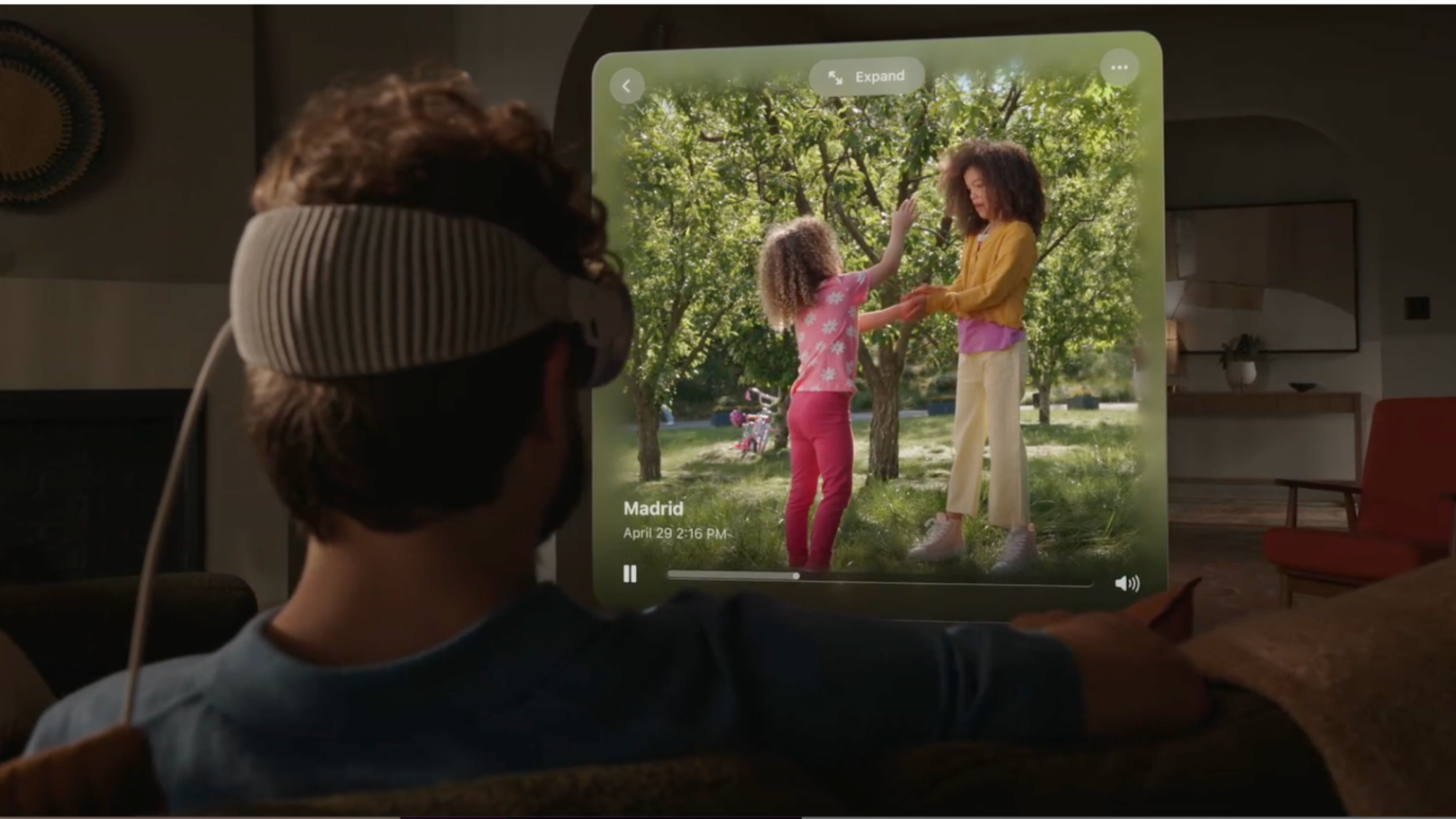 Arkadan Apple Vision Pro takan beyaz kıvırcık saçlı bir adam görülüyor.  Önüne yansıtılan ekranda iki kızın (muhtemelen kızlarının) videosunu izliyor